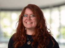 Smiling woman with long, wavy red hair and round glasses wearing a black sweater, outdoors in soft light.