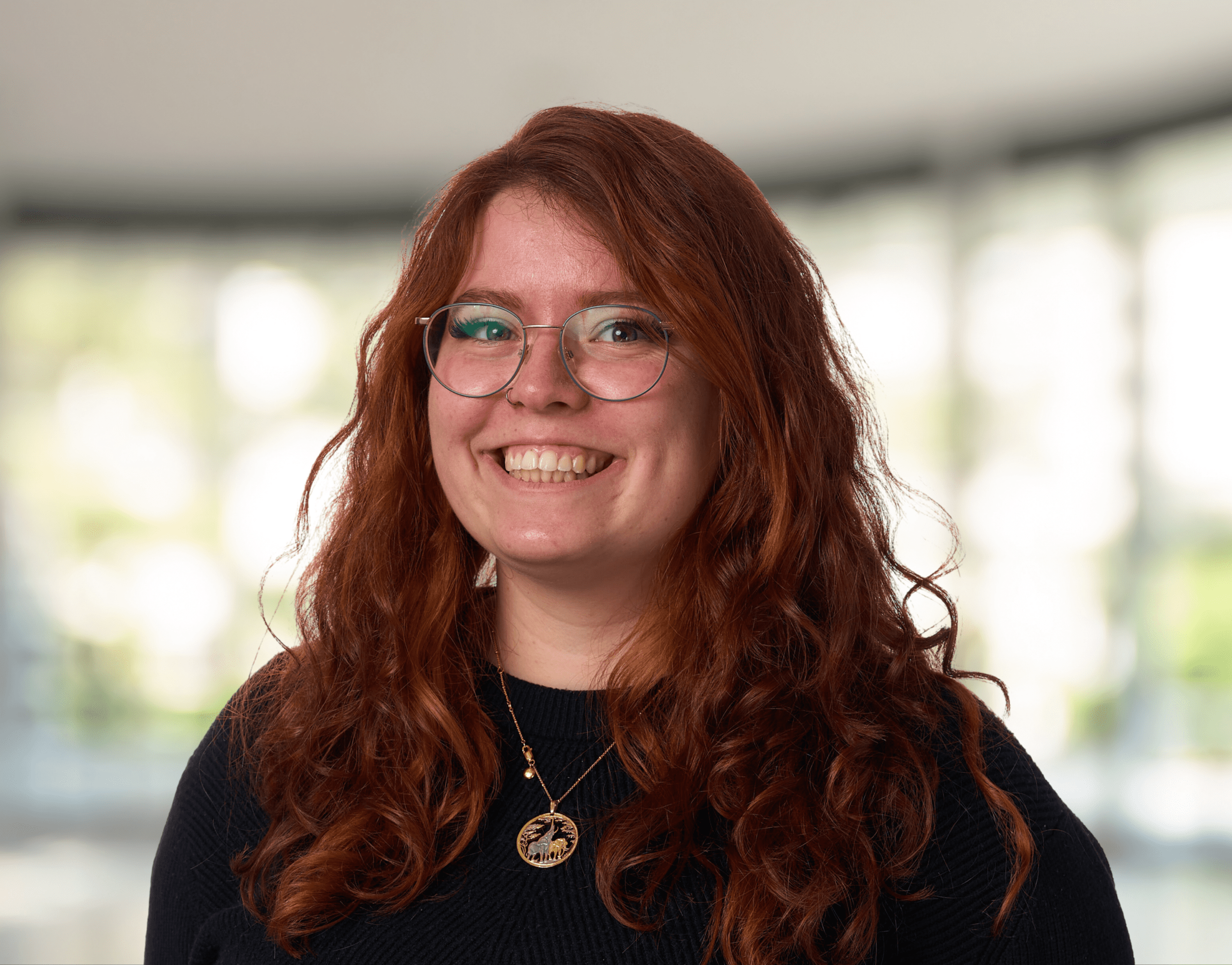 Smiling woman with long, wavy red hair and round glasses wearing a black sweater, outdoors in soft light.