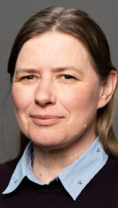 Close-up portrait of a woman with light skin and brown hair, wearing a striped collared shirt and dark sweater, smiling softly at the camera.