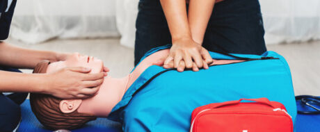 Two people perform CPR on an adult manikin: one holds the head while the other chest-compresses, with a red first aid kit nearby on a blue mat.