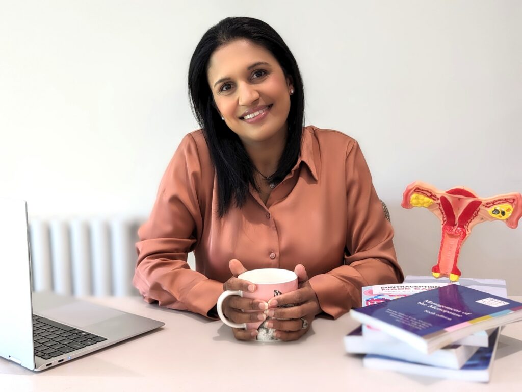 Smiling woman in a peach blouse sits at a desk, holding a pink mug, with a laptop to her left and a pelvic anatomy model on the right.