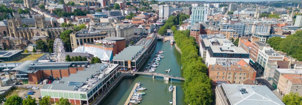 Aerial view of a city canal with boats, a pedestrian bridge, and surrounding modern buildings along the waterway.