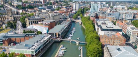 Aerial view of a city canal with boats, a pedestrian bridge, and surrounding modern buildings along the waterway.