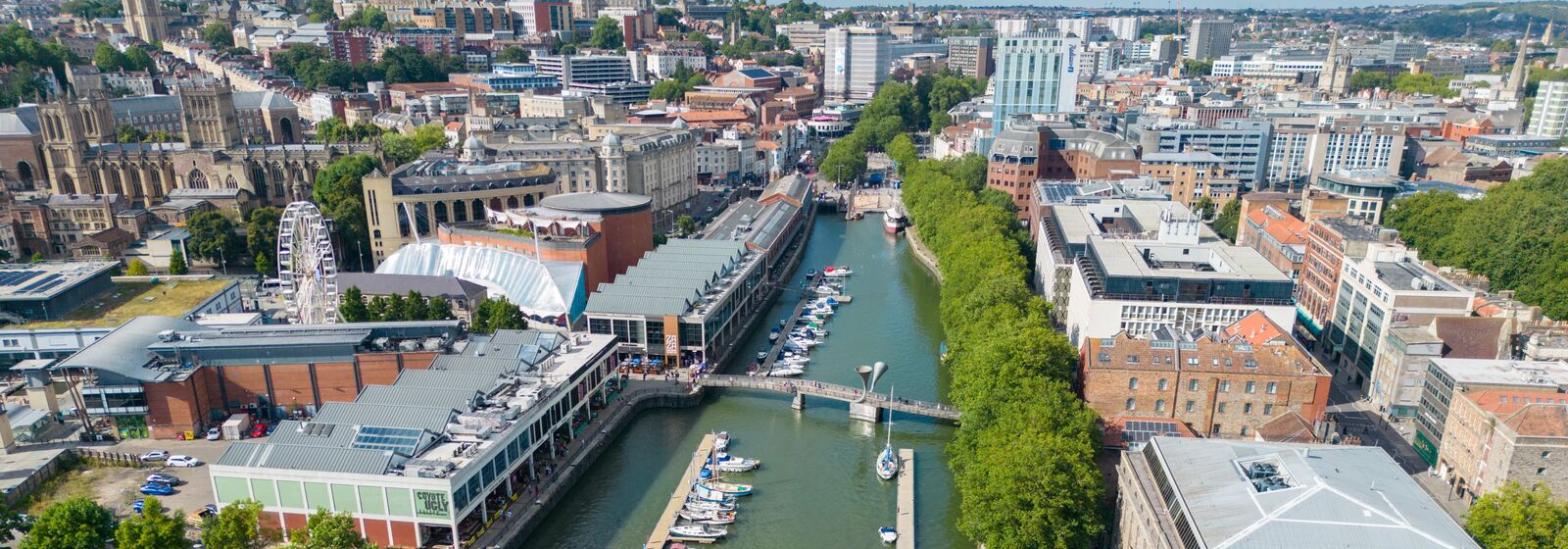 Aerial view of a city canal with boats, a pedestrian bridge, and surrounding modern buildings along the waterway.