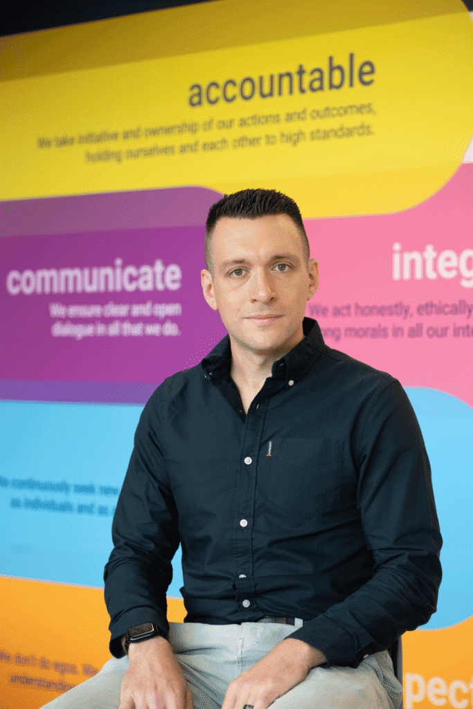 Portrait of a man in a dark shirt seated against a bright, color-blocked wall featuring the word 'accountable' and other blurred text in the background, looking at the camera.