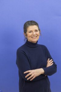 Smiling woman with dark hair in a bun, wearing a navy turtleneck, arms crossed against a blue background.