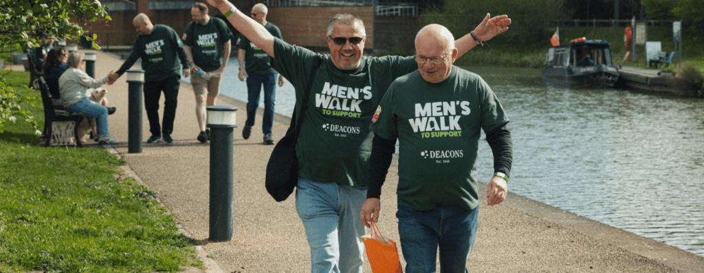 Two men in green 'Men's Walk to Support' shirts walk along a riverside path with arms raised, others follow in the background by the canal.