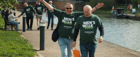 Two men in green 'Men's Walk to Support' shirts walk along a riverside path with arms raised, others follow in the background by the canal.