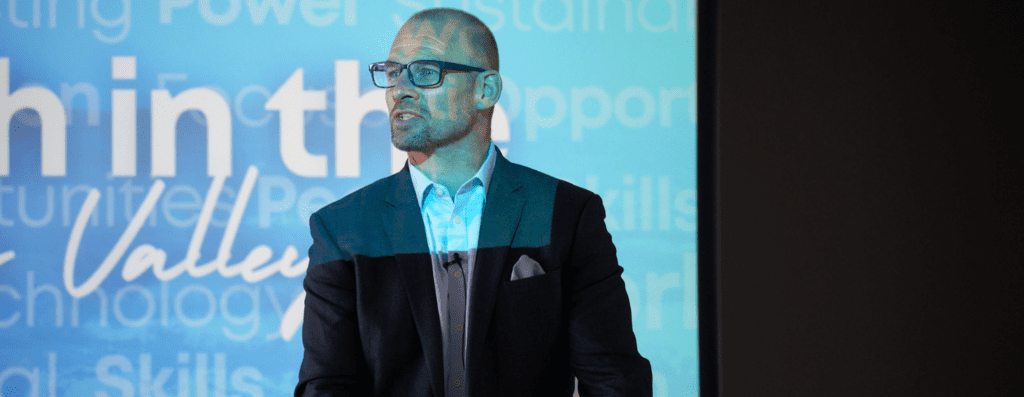 Man in a dark blazer and light blue shirt speaks at a conference, wearing glasses, with a blue tech-themed backdrop behind him.