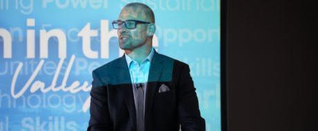 Man in a dark blazer and light blue shirt speaks at a conference, wearing glasses, with a blue tech-themed backdrop behind him.