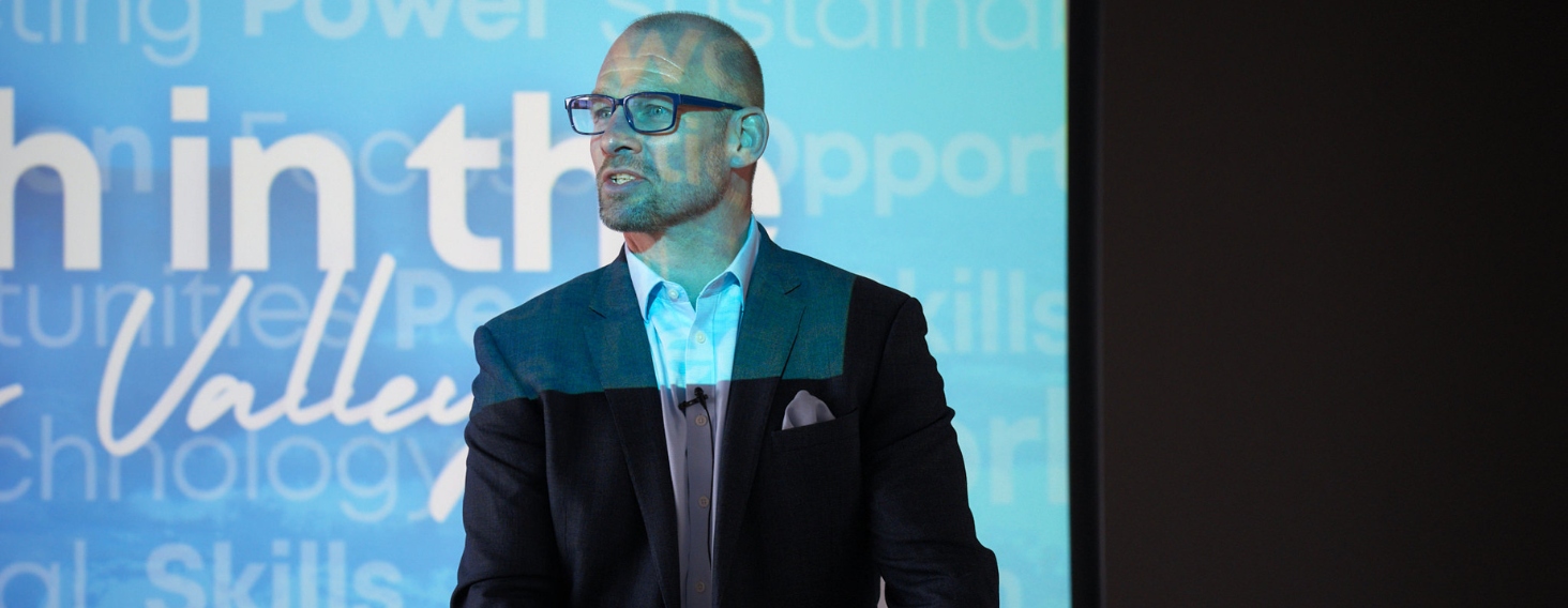Man in a dark blazer and light blue shirt speaks at a conference, wearing glasses, with a blue tech-themed backdrop behind him.