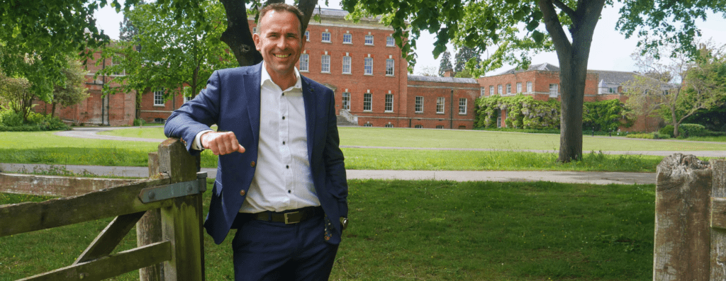 Smiling man in a navy suit leans on a wooden gate in a campus park with red brick buildings in the background.