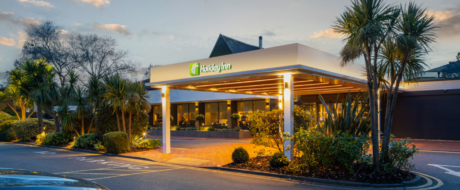 Exterior of a Holiday Inn hotel at dusk with a lit entrance, palm trees, and manicured landscaping.