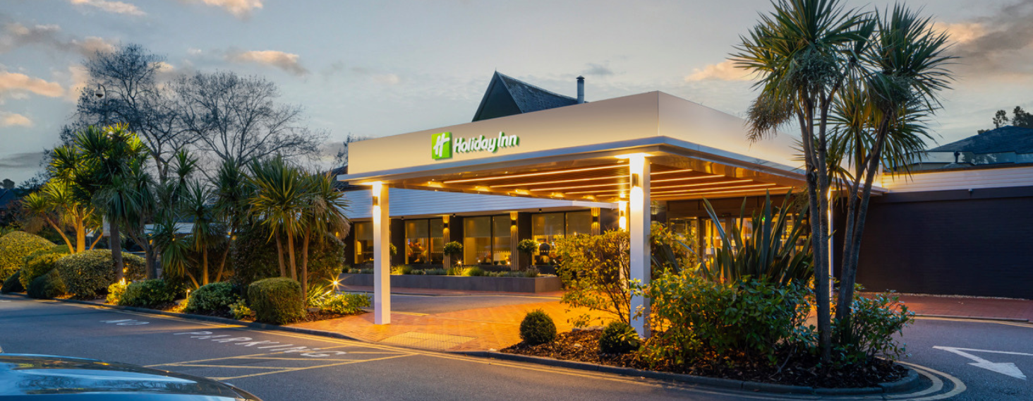 Exterior of a Holiday Inn hotel at dusk with a lit entrance, palm trees, and manicured landscaping.