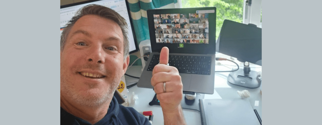 Smiling man in a home office giving a thumbs-up beside a laptop showing a Zoom grid of participants on screen.