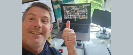 Smiling man in a home office giving a thumbs-up beside a laptop showing a Zoom grid of participants on screen.