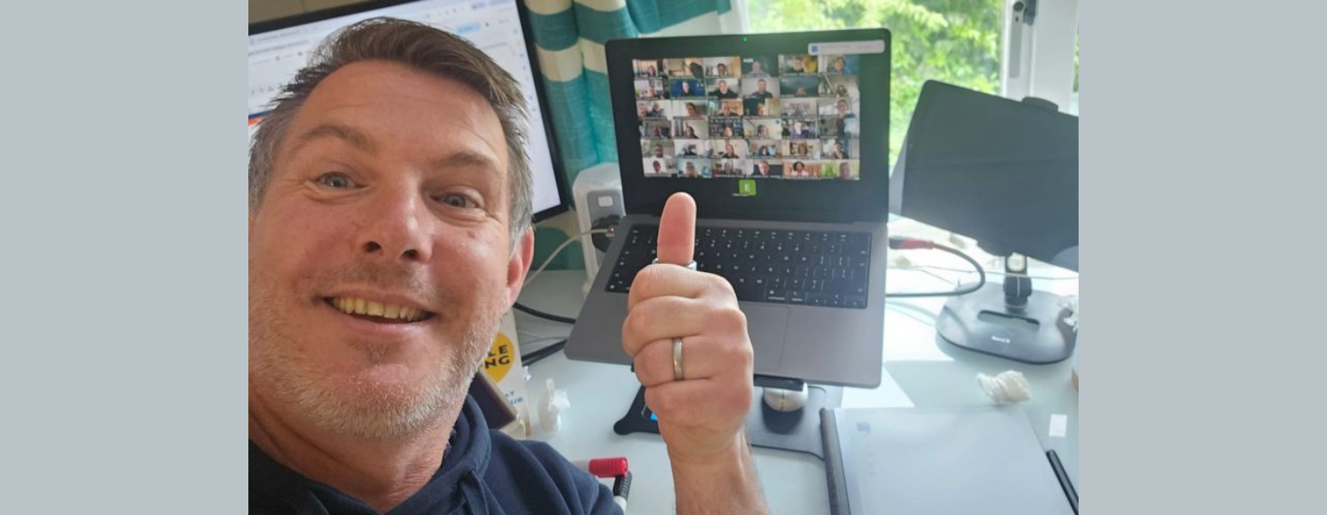 Smiling man in a home office giving a thumbs-up beside a laptop showing a Zoom grid of participants on screen.