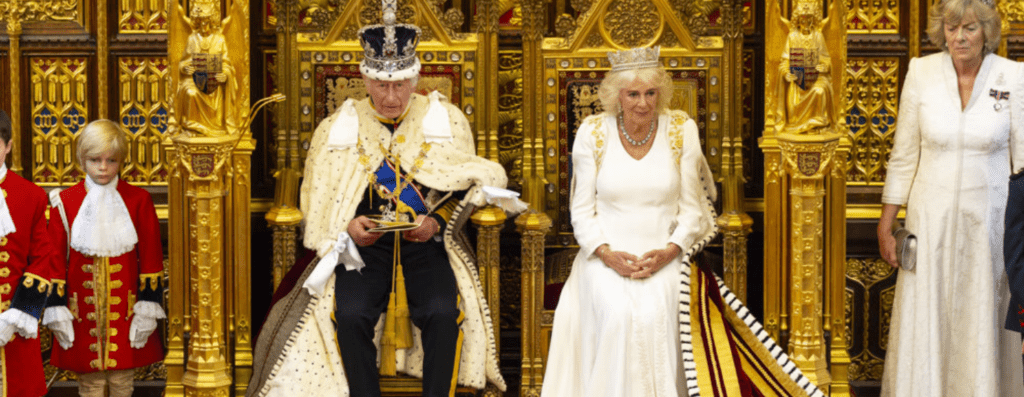 King and queen sit on ornate gold thrones during a ceremonial event, with attendants in red uniforms beside them.