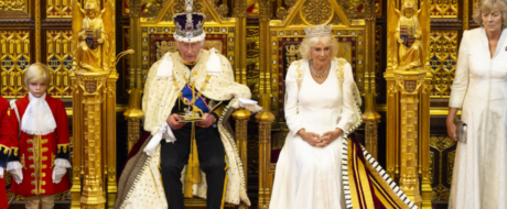 King and queen sit on ornate gold thrones during a ceremonial event, with attendants in red uniforms beside them.