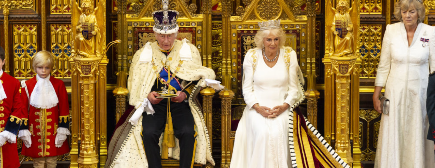 King and queen sit on ornate gold thrones during a ceremonial event, with attendants in red uniforms beside them.