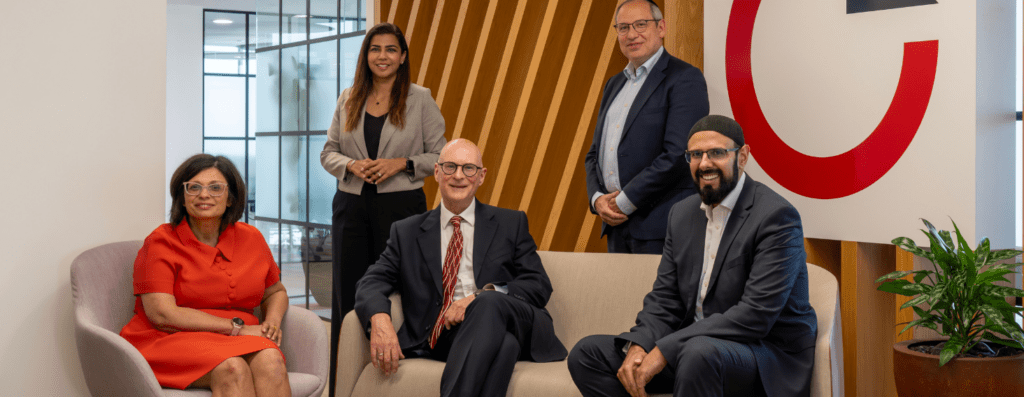 Five professionals posing in a modern office lobby with a wooden slat wall and a large red logo in the background.