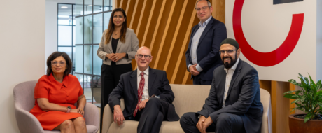Five professionals posing in a modern office lobby with a wooden slat wall and a large red logo in the background.