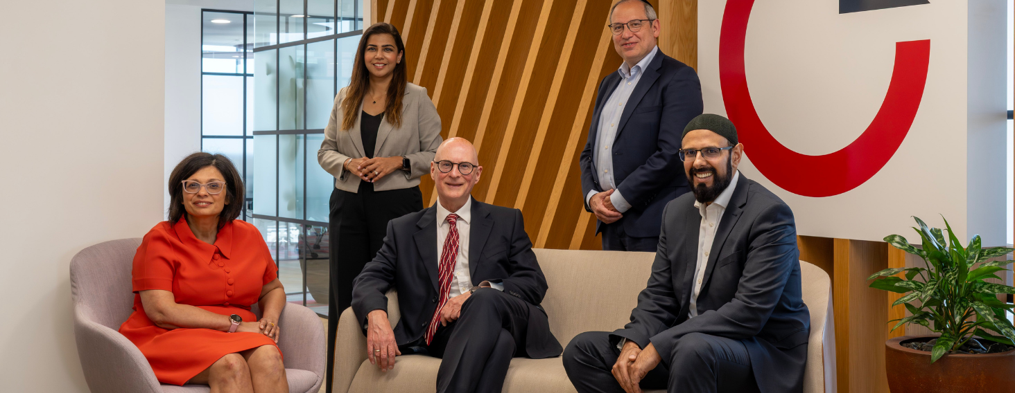 Five professionals posing in a modern office lobby with a wooden slat wall and a large red logo in the background.