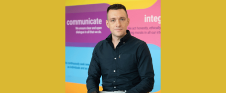 Portrait of a man in a dark blue shirt seated against a bright, multi-colored background featuring the word 'communicate'.