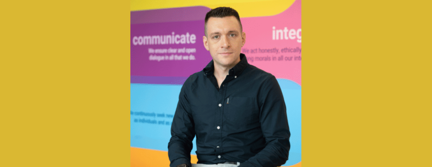 Portrait of a man in a dark blue shirt seated against a bright, multi-colored background featuring the word 'communicate'.
