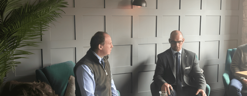 Two men in a business setting sit facing each other in a modern lounge, one in a vest and the other in a suit with glasses, discussing.