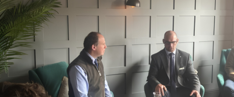Two men in a business setting sit facing each other in a modern lounge, one in a vest and the other in a suit with glasses, discussing.