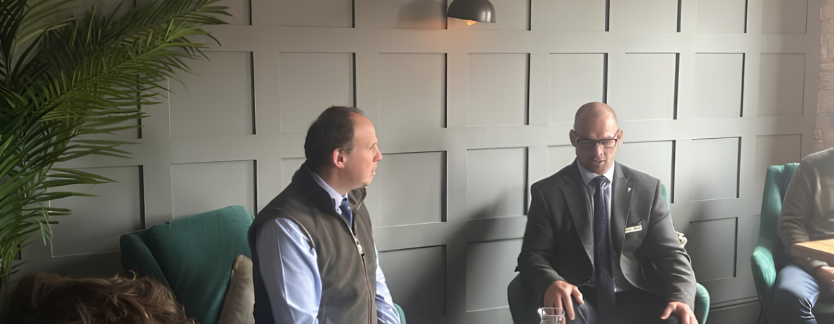 Two men in a business setting sit facing each other in a modern lounge, one in a vest and the other in a suit with glasses, discussing.