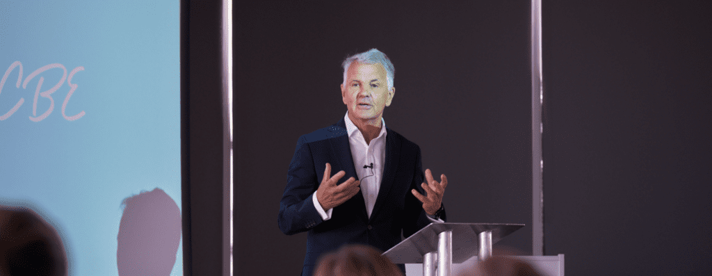 Male speaker in a dark suit delivering a presentation at a clear podium with a mic, addressing an audience in a conference room