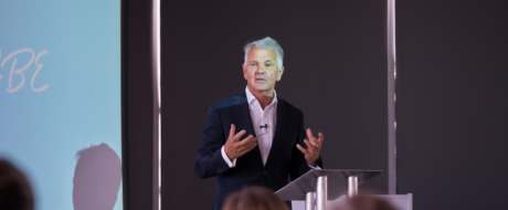 Male speaker in a dark suit delivering a presentation at a clear podium with a mic, addressing an audience in a conference room