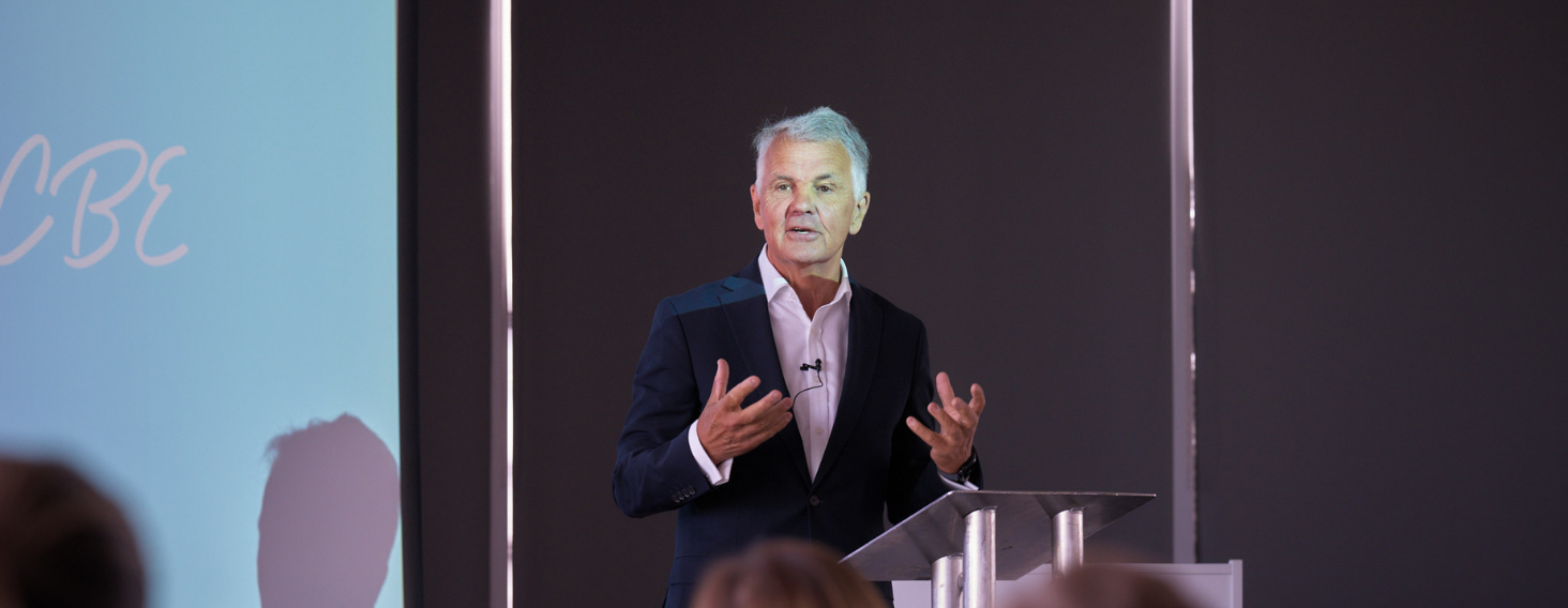 Male speaker in a dark suit delivering a presentation at a clear podium with a mic, addressing an audience in a conference room