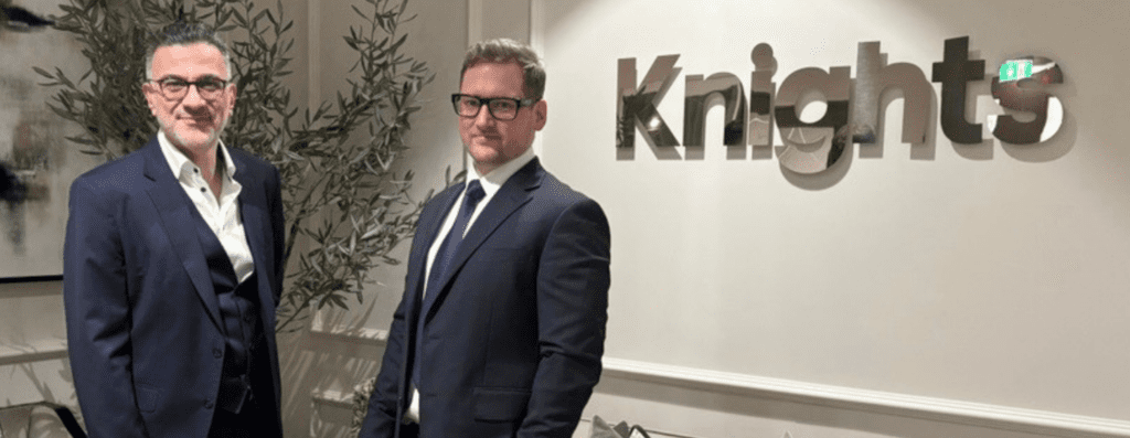 Two men in dark suits stand side by side in an office lobby with a large knig hts sign on the wall behind them. A tall plant sits to their left.