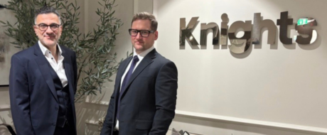 Two men in dark suits stand side by side in an office lobby with a large knig hts sign on the wall behind them. A tall plant sits to their left.