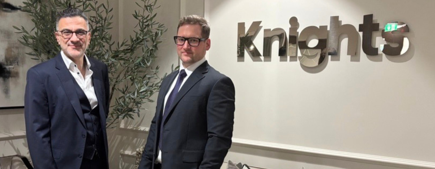 Two men in dark suits stand side by side in an office lobby with a large knig hts sign on the wall behind them. A tall plant sits to their left.