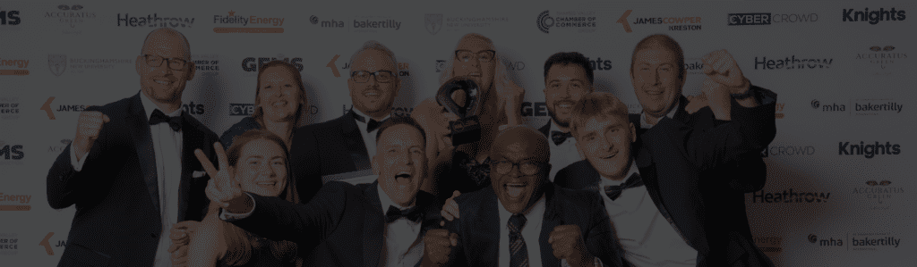 Group of people in tuxedos posing and cheering at an awards event, with a trophy and sponsor logos backdrop.