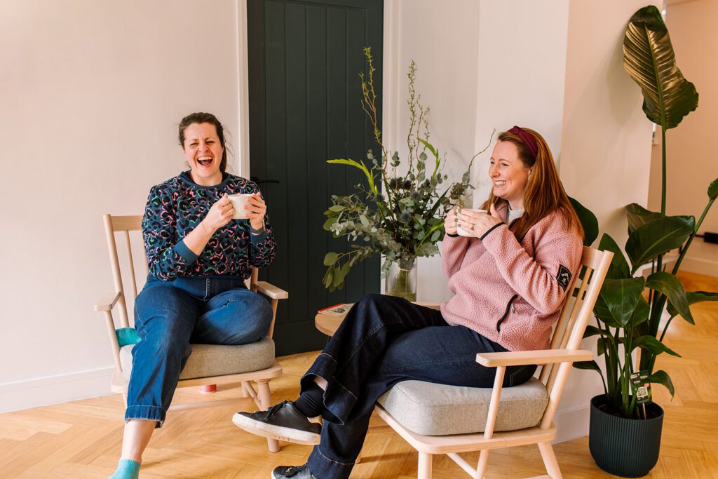 Two women sit in wooden chairs facing each other, laughing and holding mugs in a bright, cozy room.