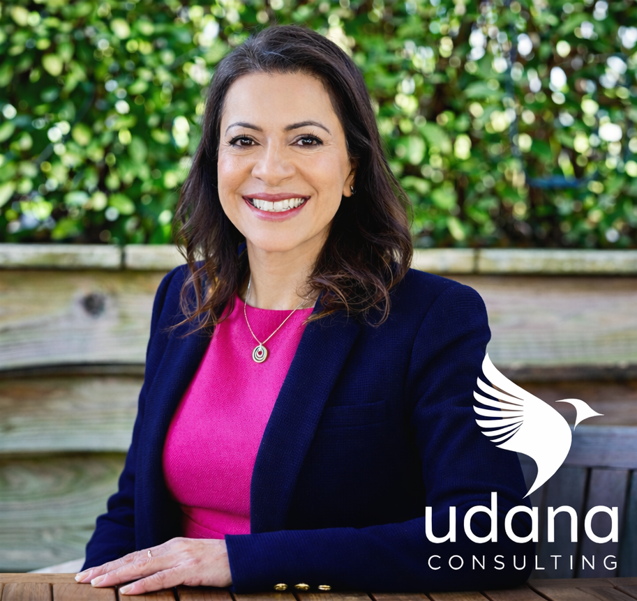 Smiling woman in a pink top and navy blazer sits on a wooden bench outdoors, with the Udana Consulting logo in the lower right corner.