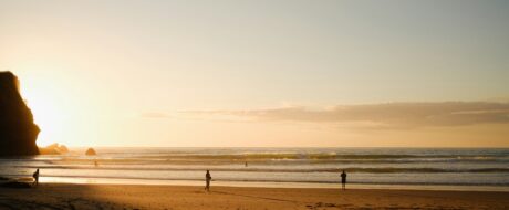 Sunset beach scene with a rocky cliff on the left and a few silhouetted people along the shore.