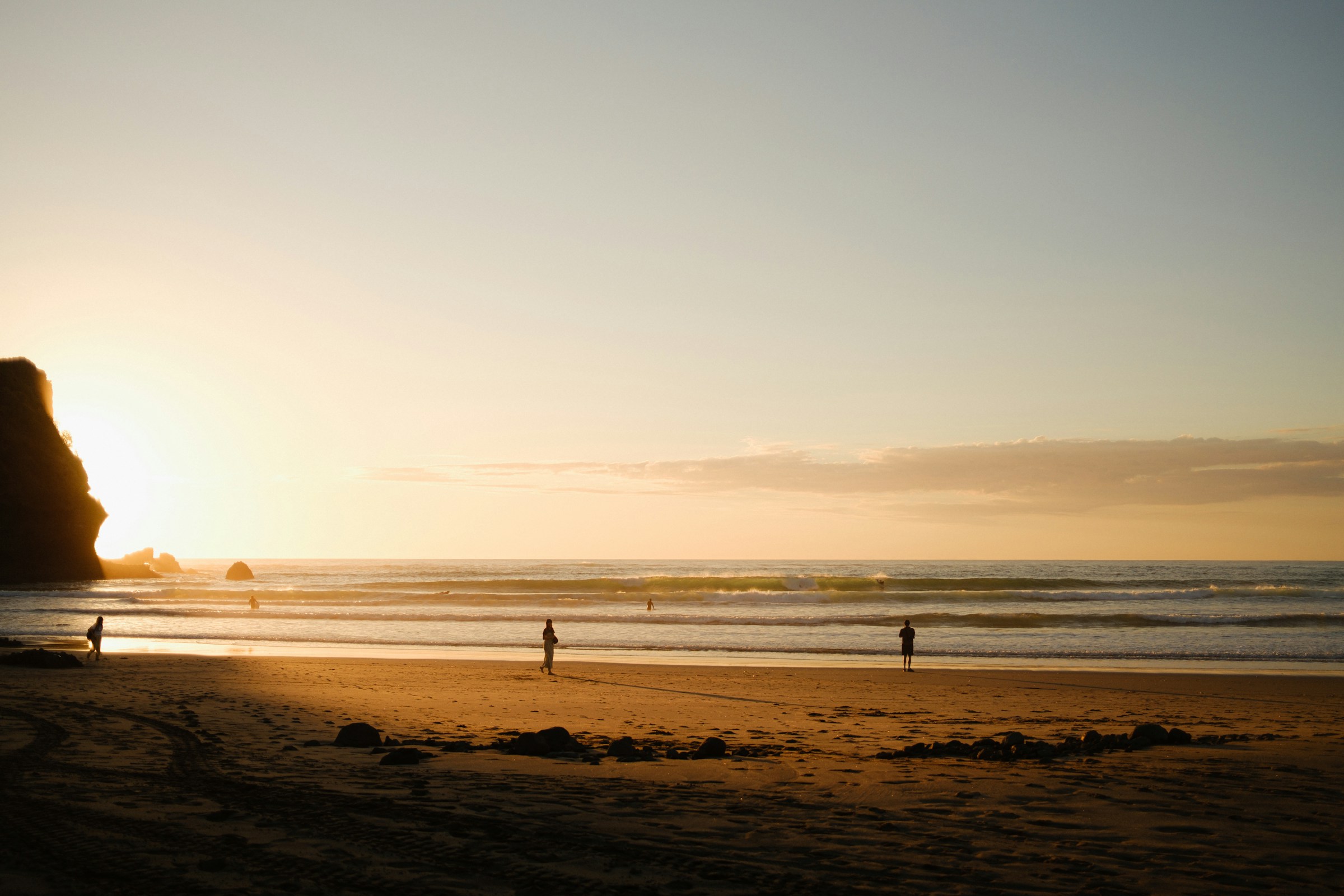 Sunset beach scene with a rocky cliff on the left and a few silhouetted people along the shore.