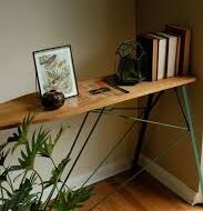 Wooden desk with metal legs in a corner, with books, a framed photo, and a small plant on the surface; a large plant sits in the foreground.