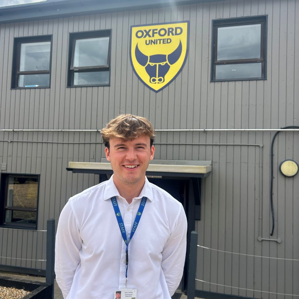 Young man in a white shirt with a staff lanyard smiles at the camera, standing in front of a gray building with the Oxford United crest.",