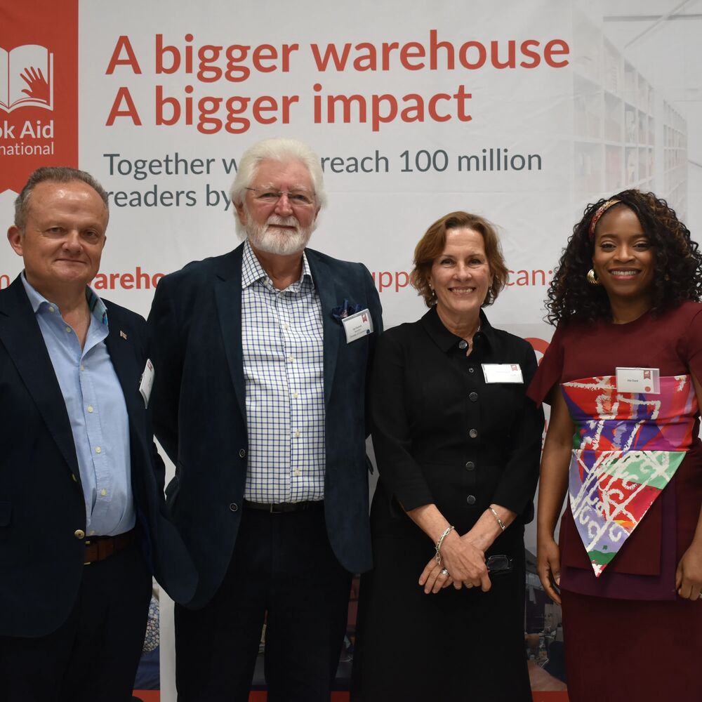 Four professionally dressed individuals pose together in front of a banner that says 'A bigger warehouse A bigger impact'.