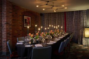 Long dining table set for a formal dinner with candles, flowers, glassware, and a brick-walled room with artistic decor.