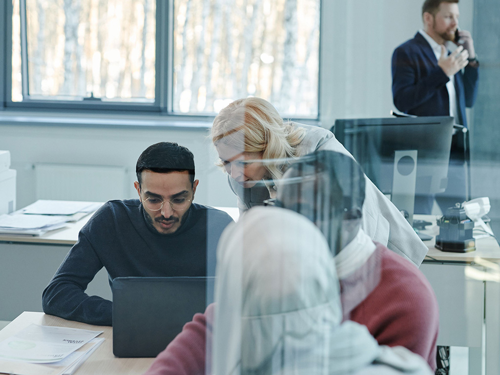 Team of coworkers collaborating at a desk with laptops; two people in the foreground lean in as others discuss.