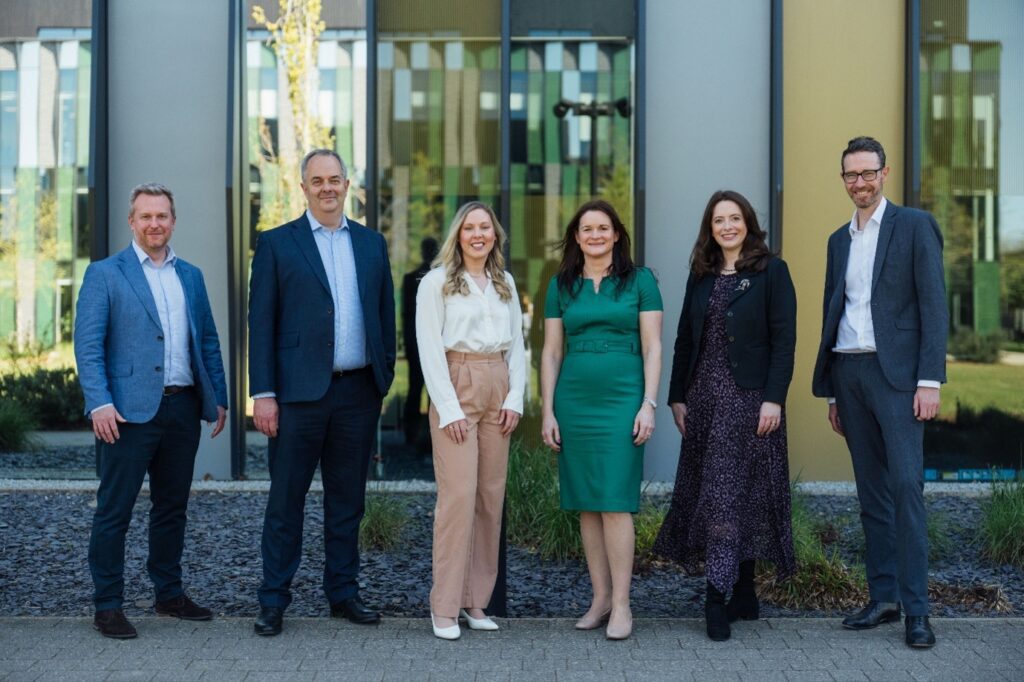 Six professionals in business attire stand outside a modern glass-front building, posing for a group photo on a paved walkway with landscaping nearby.
