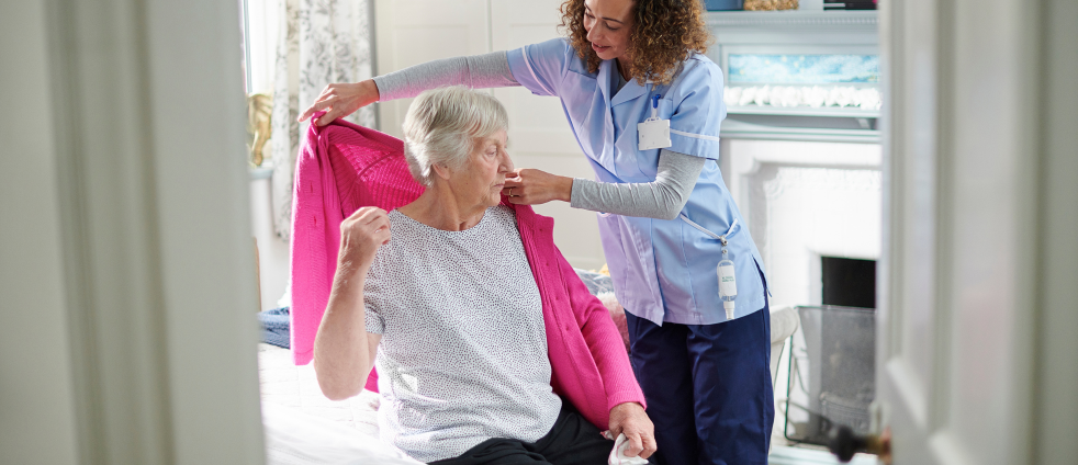 Nurse in blue scrubs helps an elderly woman put on a pink cardigan beside a bed or in a care setting.